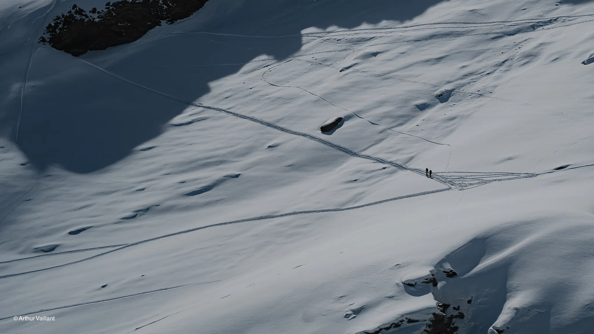Two skiers are making their way across a snow-covered Alpine landscape, bathed in sunlight.