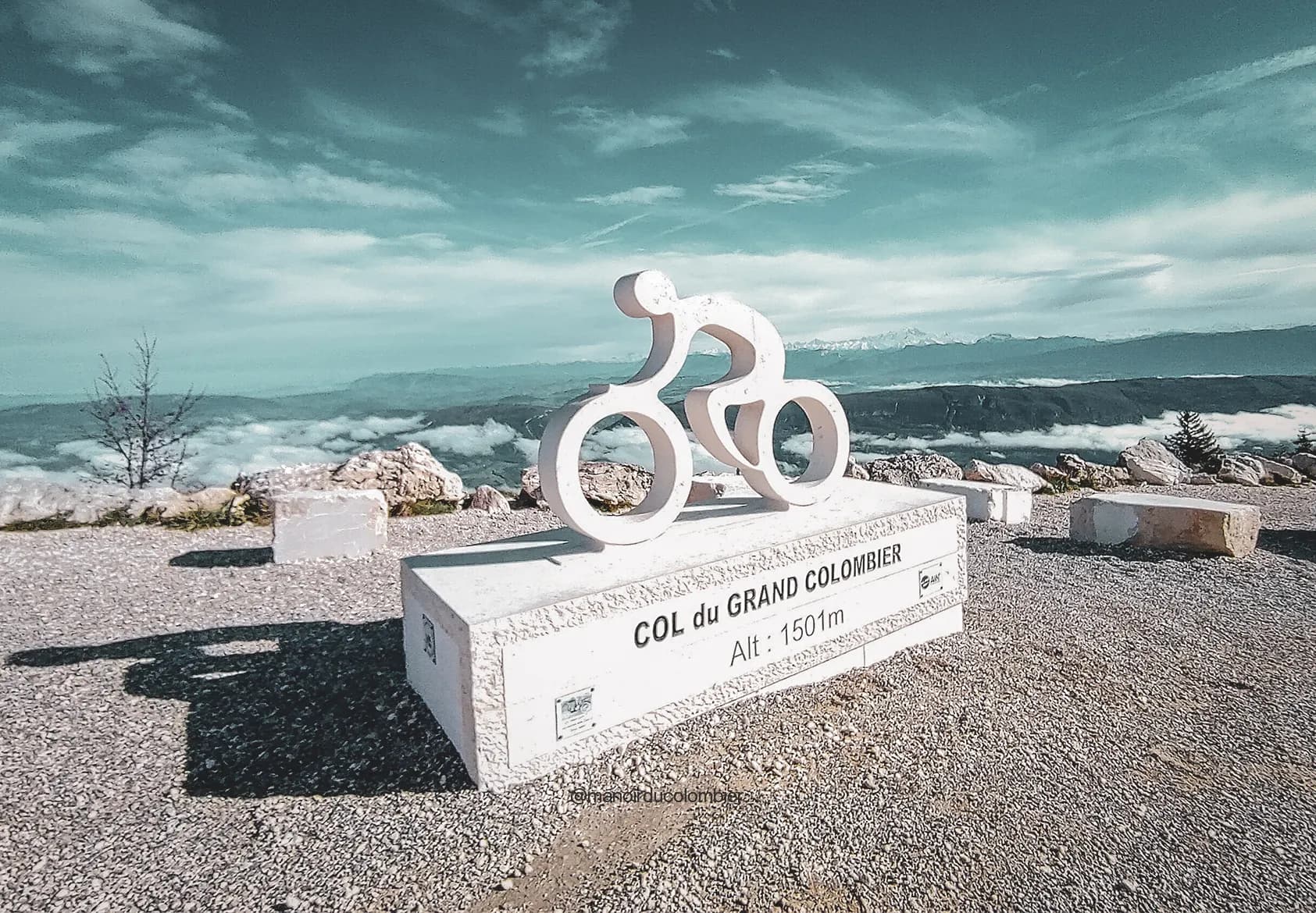 Vue panoramique au Col du Grand Colombier, avec une sculpture de cycliste sur fond de montagnes.