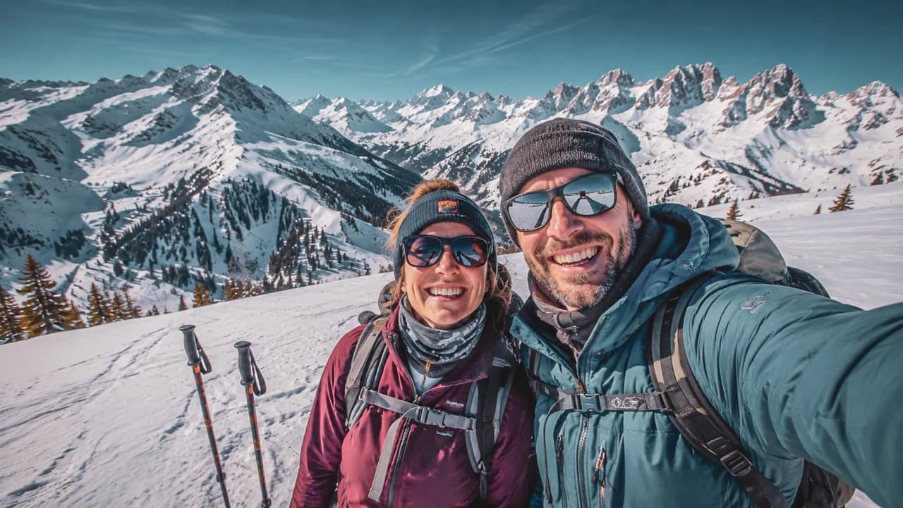 A smiling couple on skis, set against snow-capped mountains under a blue sky, in a mountain setting.