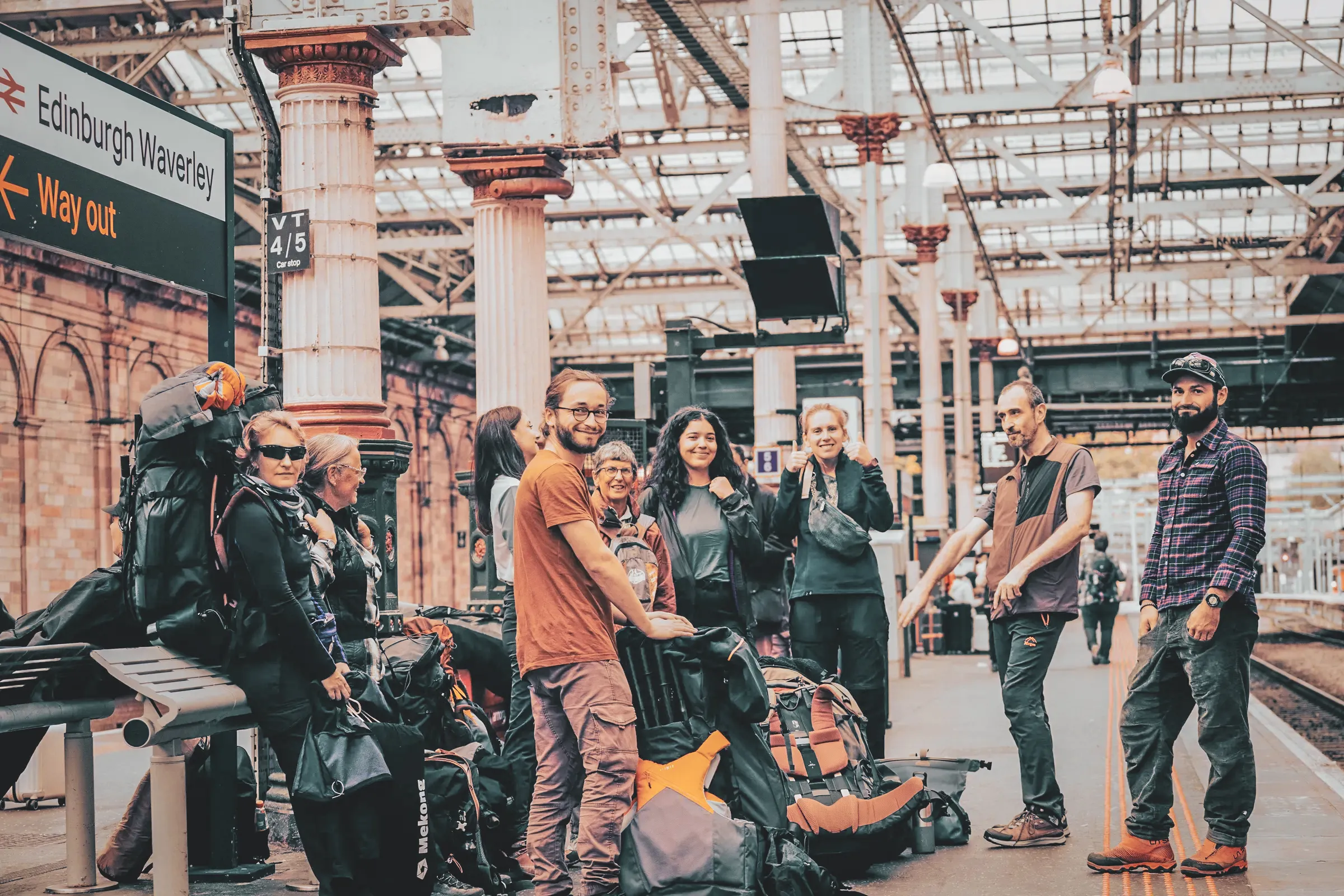 Groepen lachende wandelaars met hun rugzakken op het station van Edinburgh, klaar voor avontuur.