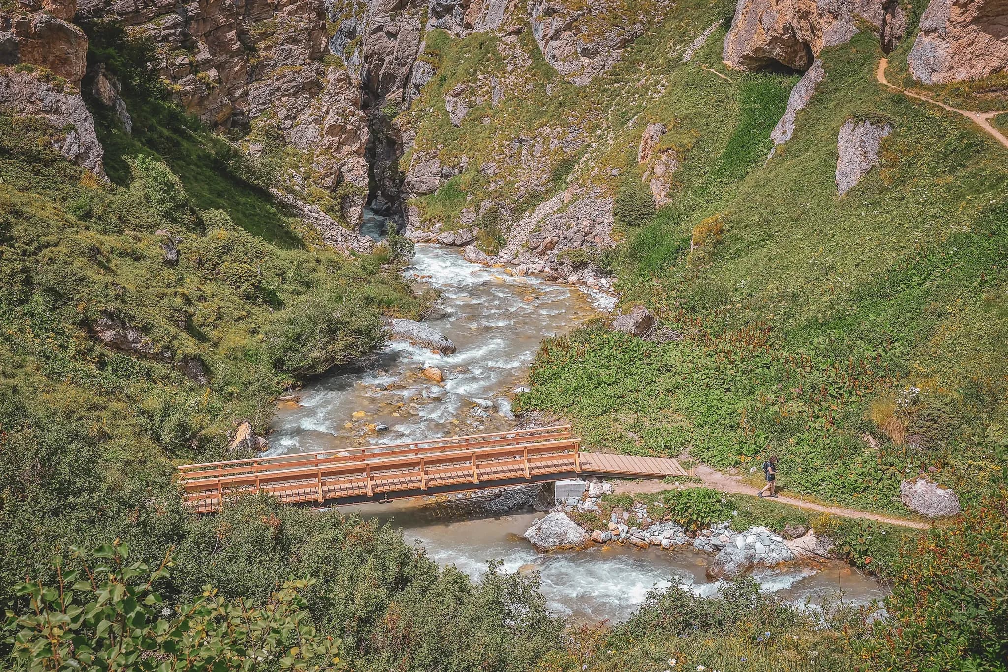 A wooden bridge crosses a lively river, surrounded by greenery and majestic Alpine scenery.