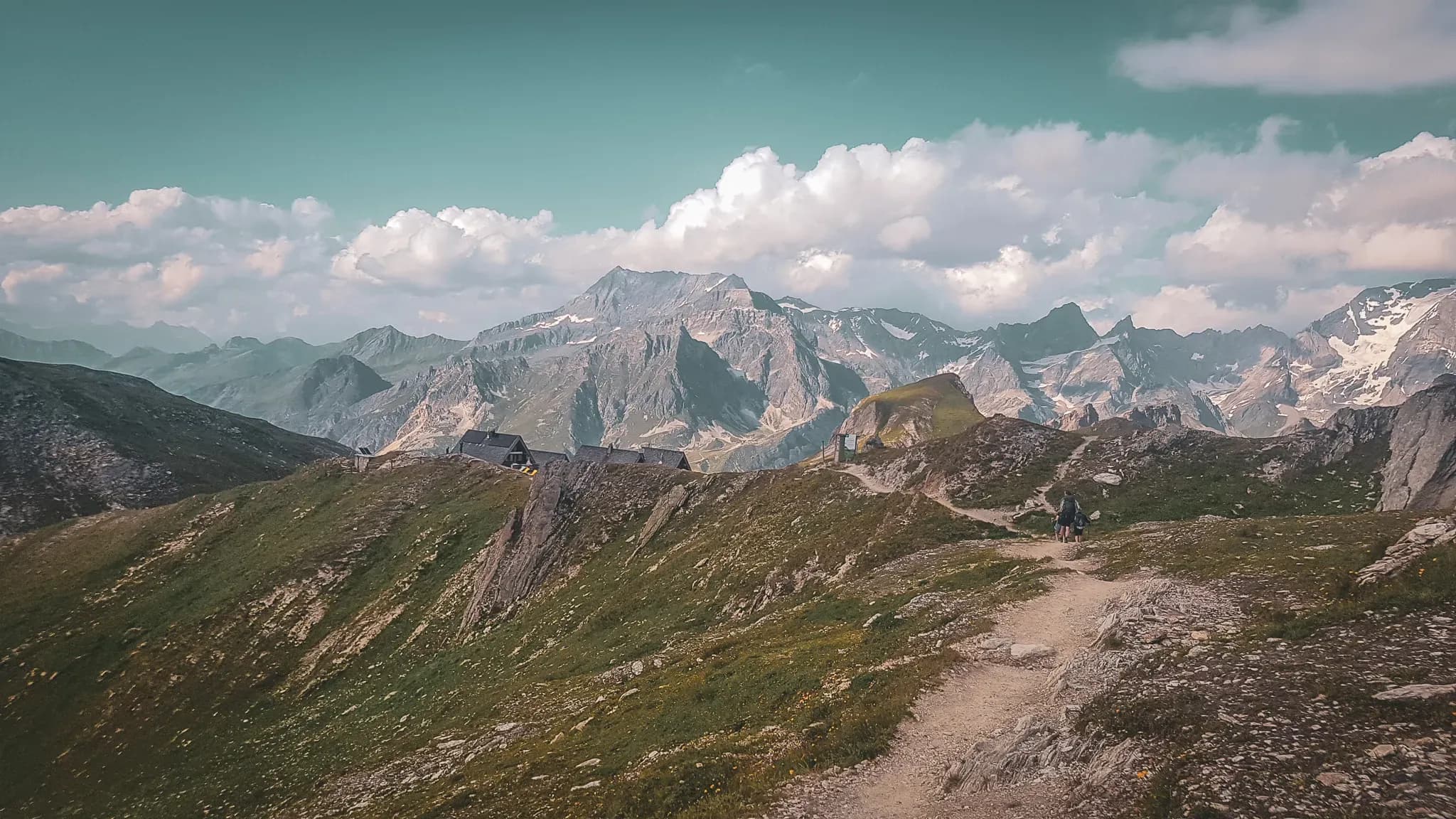 A mountain trail with huts, surrounded by majestic Alpine peaks and blue skies.