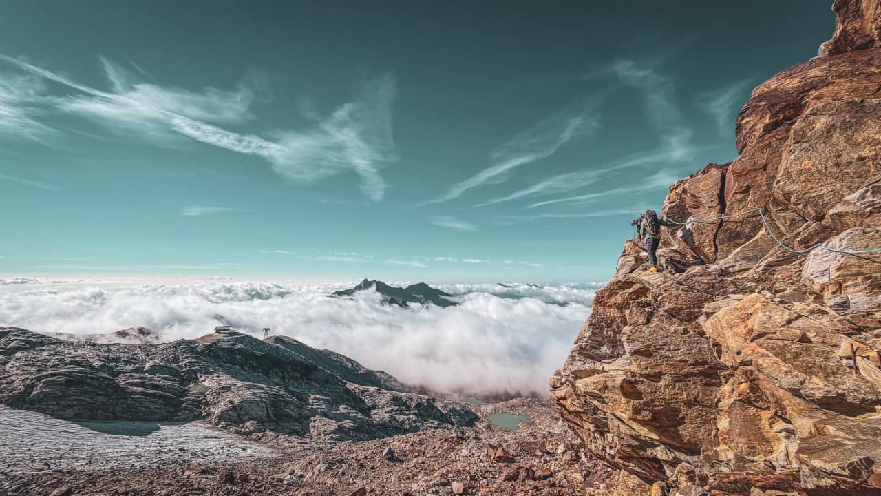 A mountaineer crossing a rock ledge overlooking an ocean of clouds beneath a stunning blue sky.