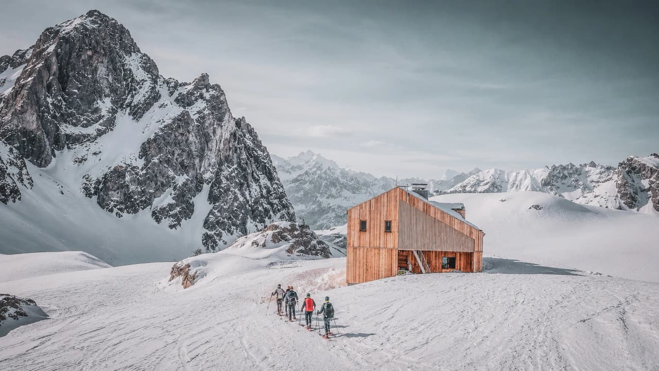 A group of explorers on snowshoes, making their way to a wooden Mountain hut surrounded by snow-capped peaks.
