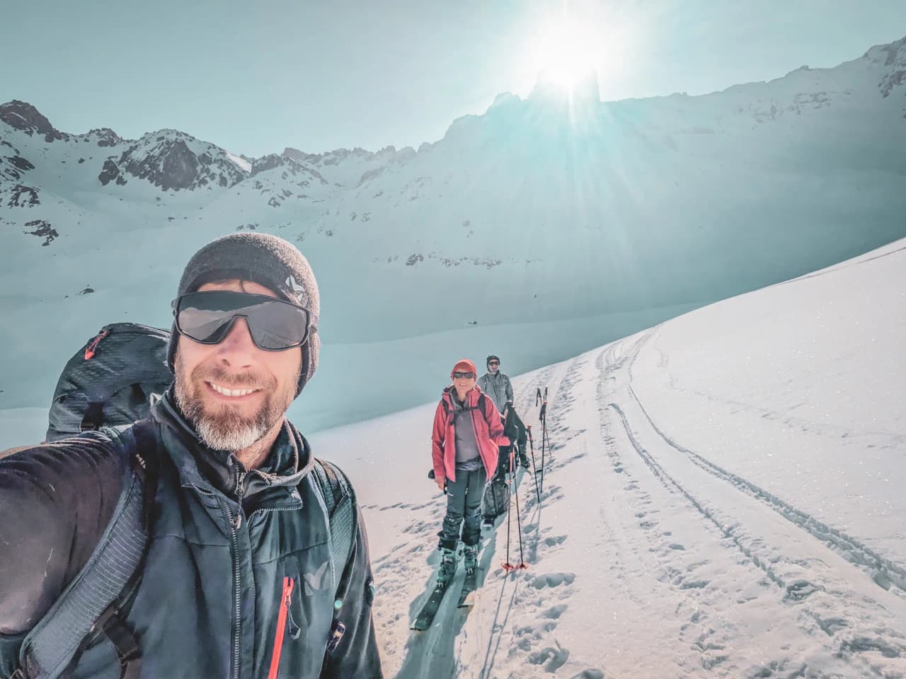 Three skiers on a snow-covered Alpine landscape, with the sun shining brightly in the background.