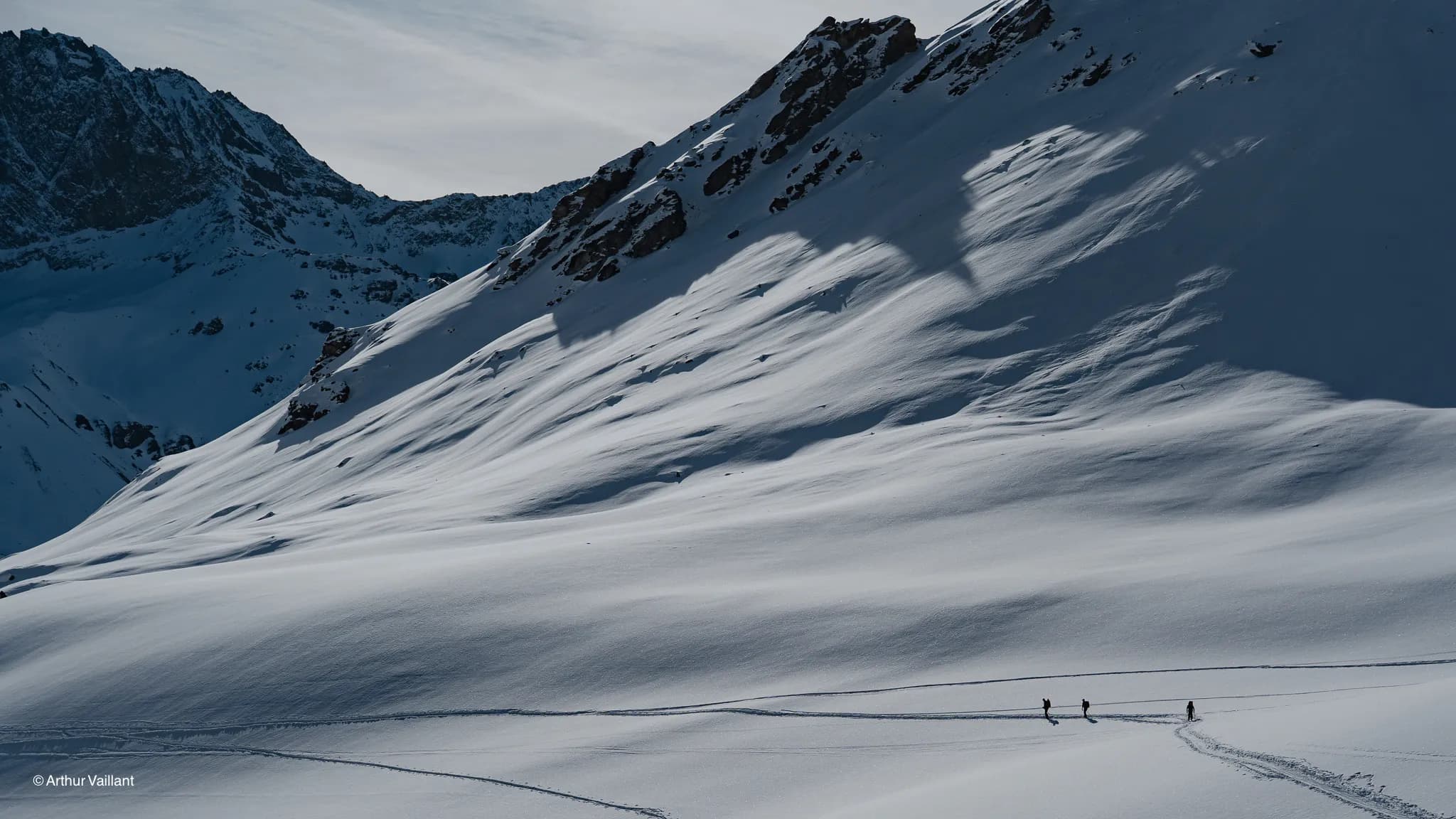 Three adventurers make their way through a pristine alpine landscape, amidst majestic mountains and glistening snow.