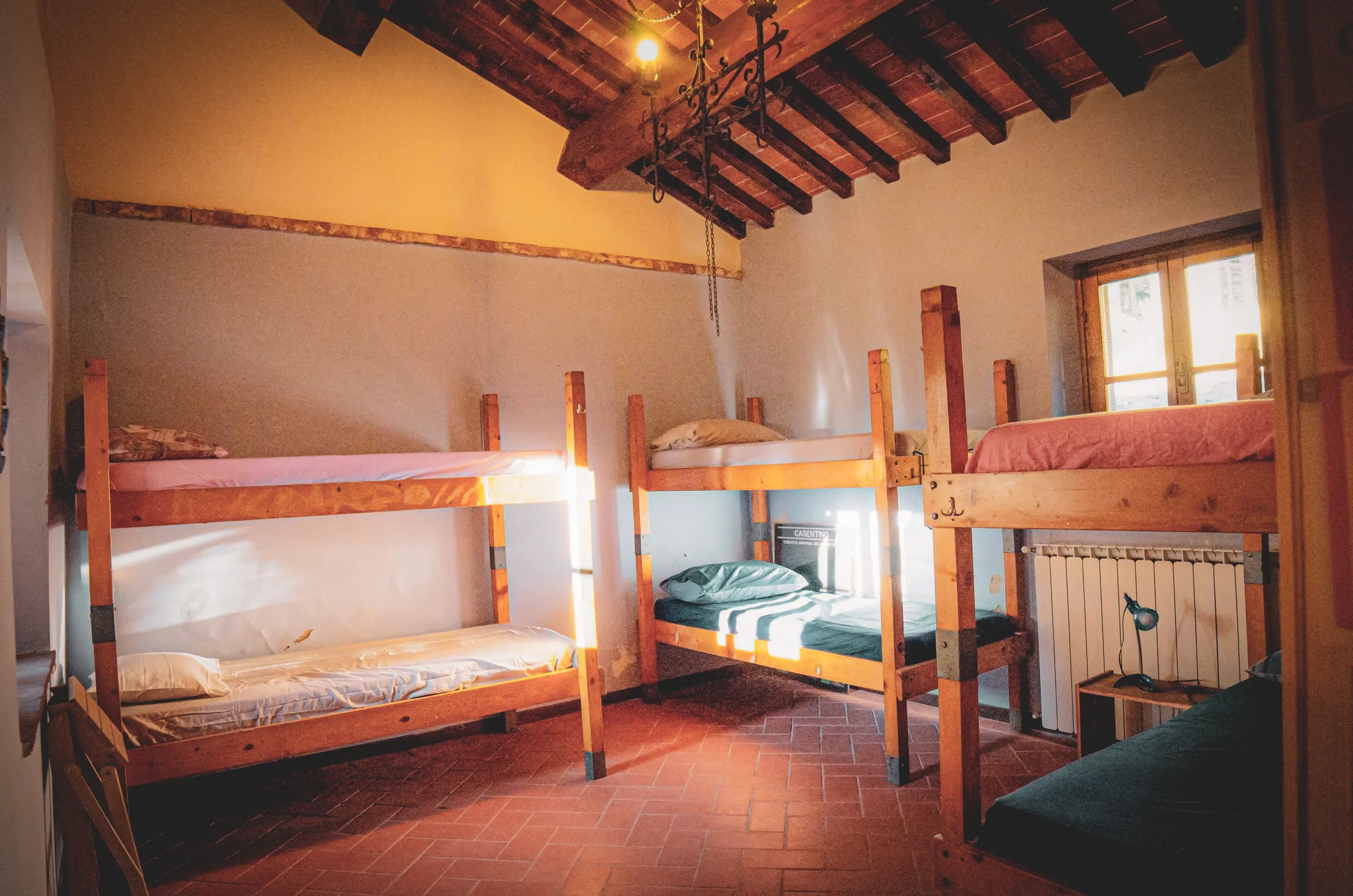 Une chambre avec des lits superposés en bois, offrant un espace accueillant et fonctionnel. Le sol est en carrelage rouge et les murs sont peints dans des tons neutres. Au fond, une fenêtre laisse entrer la lumière naturelle