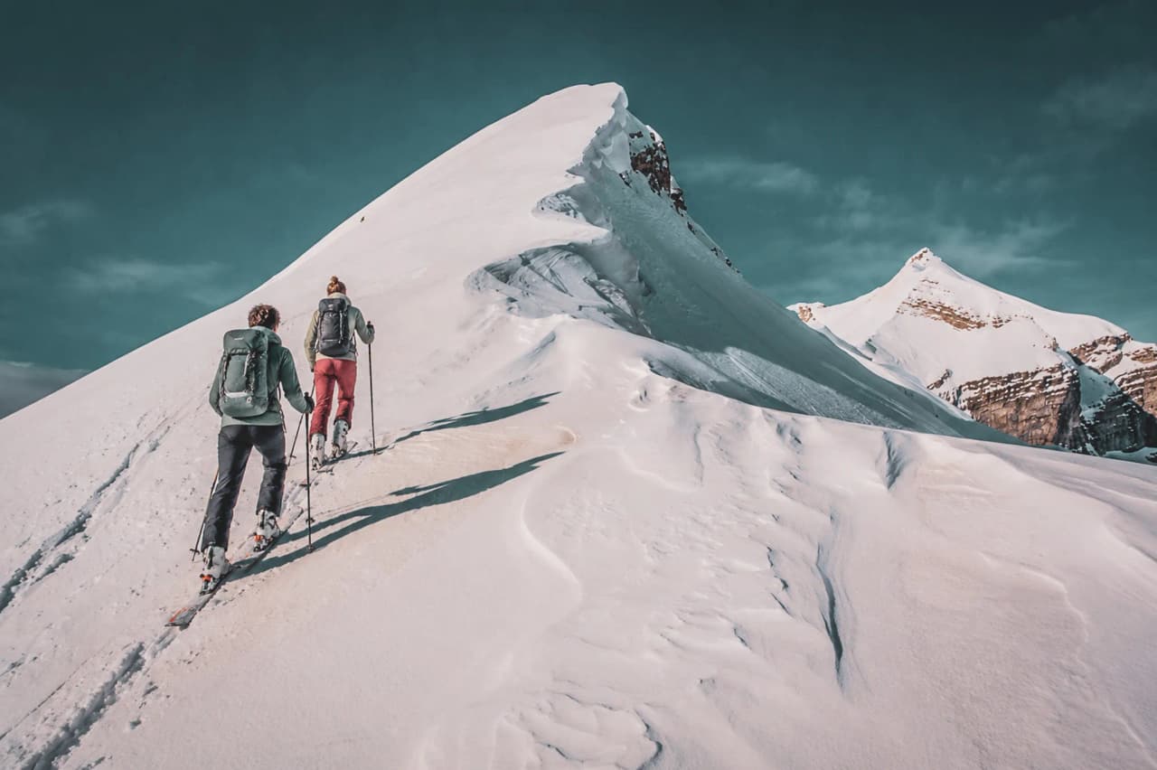 Two skiers are climbing a snow-covered ridge, surrounded by the majestic Écrins mountains.
