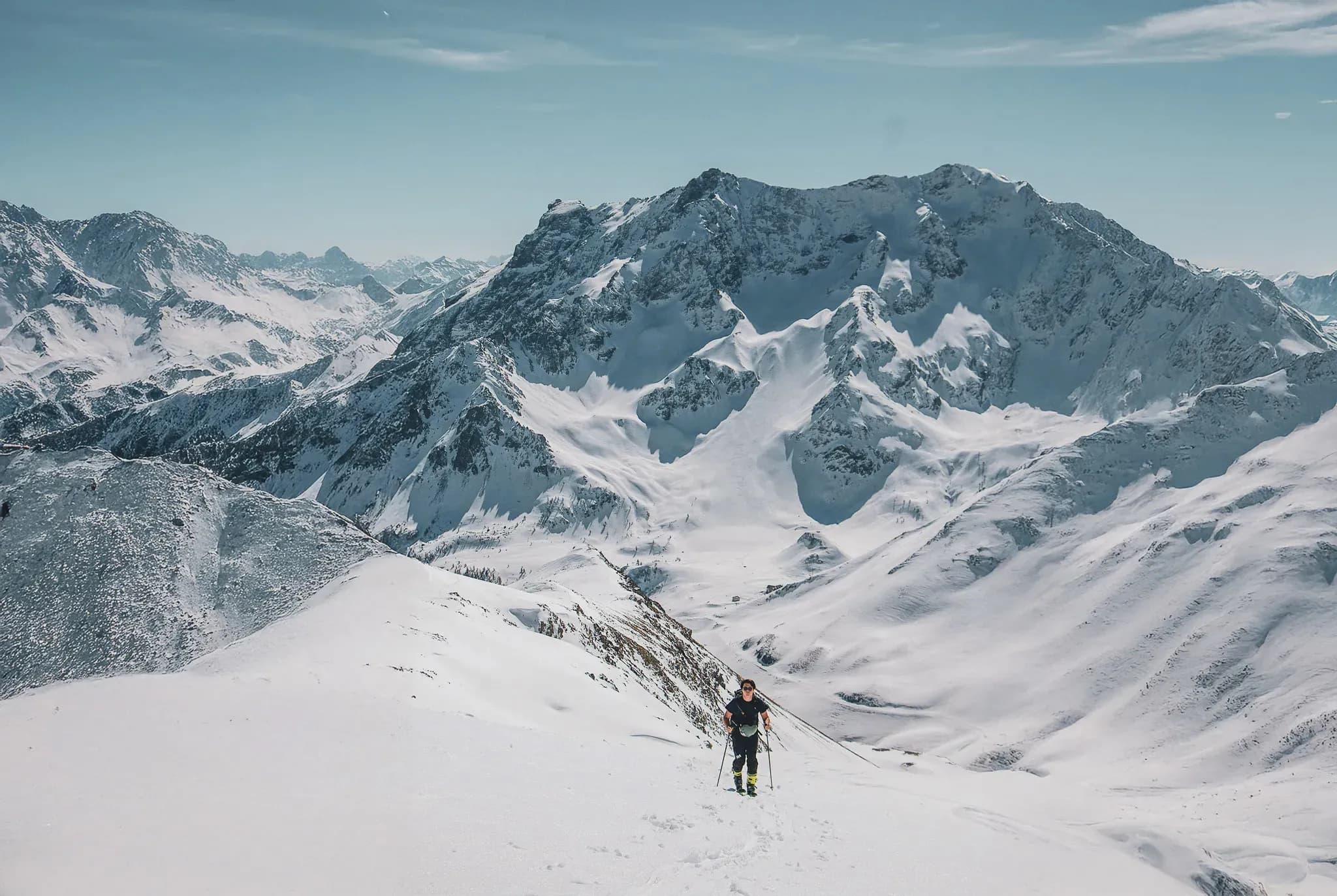 A lone skier climbing majestic snow-capped peaks under a brilliant blue sky.