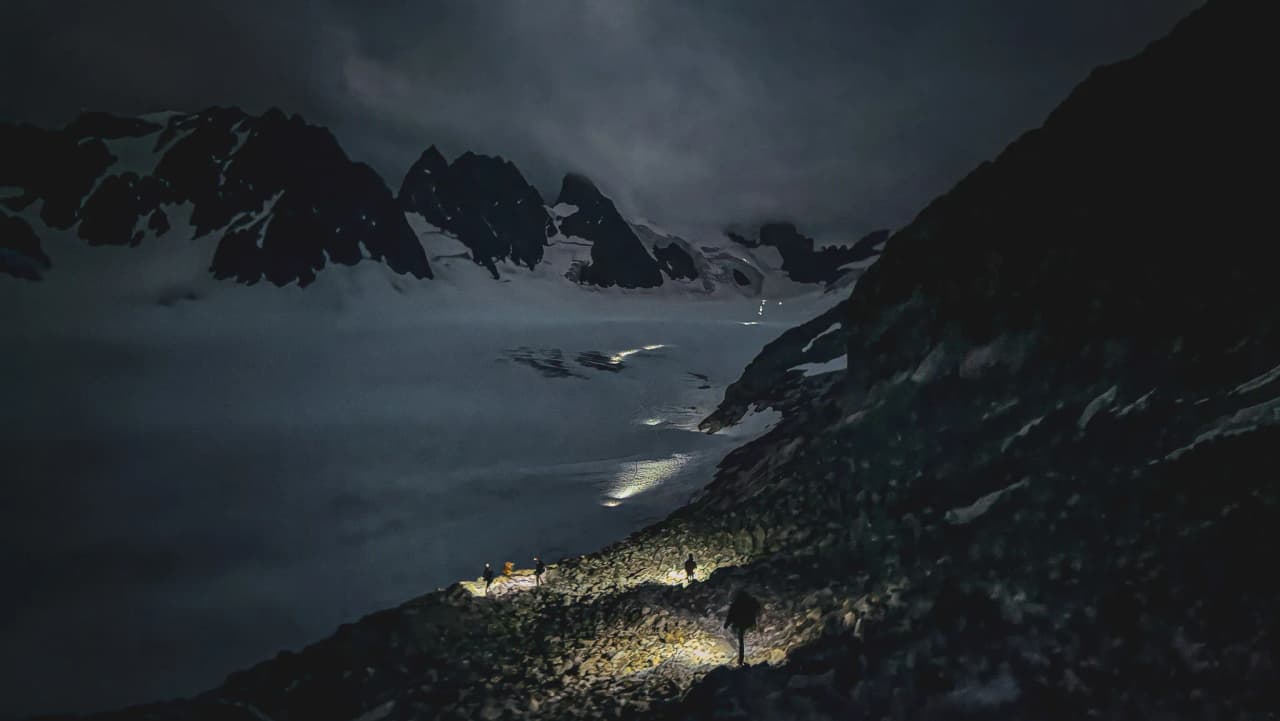 Une expédition nocturne en montagne, avec des alpinistes illuminés par des frontales sur un chemin rocailleux, entourés de majestueux sommets enneigés.