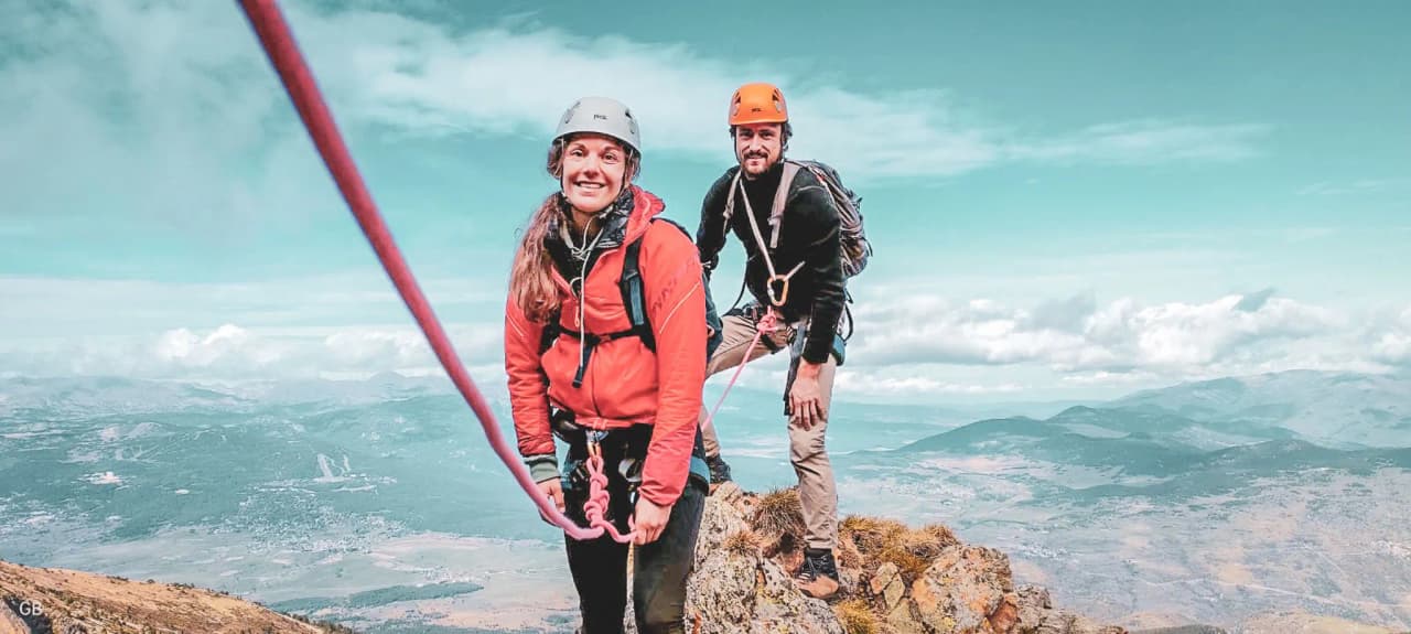 Duo d'alpinistes souriants au sommet des Pyrénées, avec un panorama spectaculaire en arrière-plan.