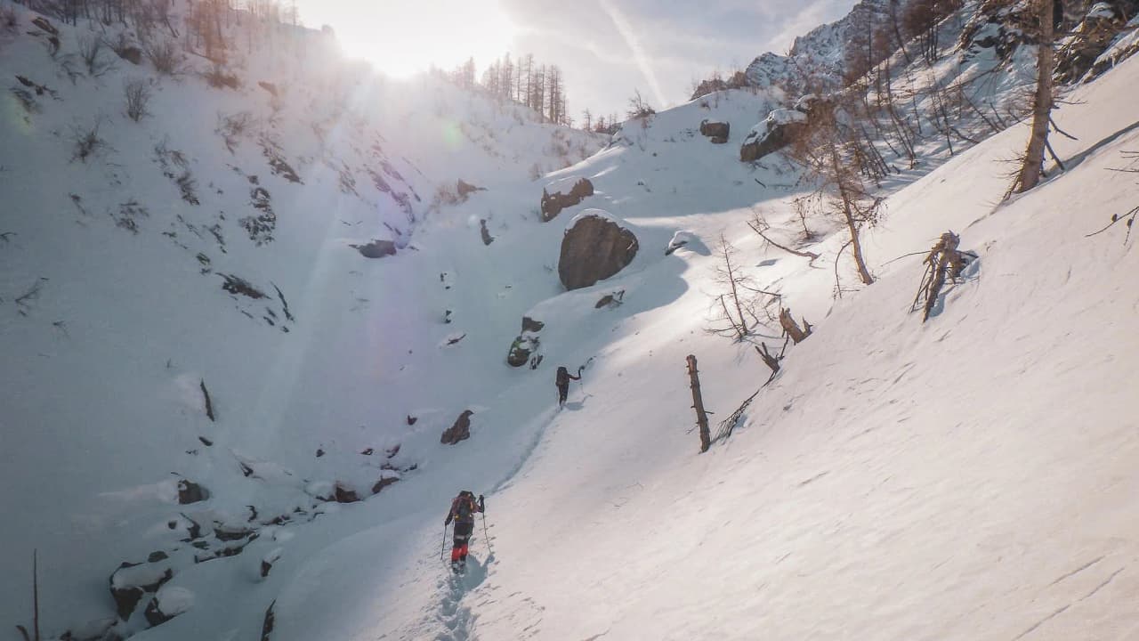 Two hikers make their way up a snow-covered slope, surrounded by majestic mountains in brilliant sunshine.
