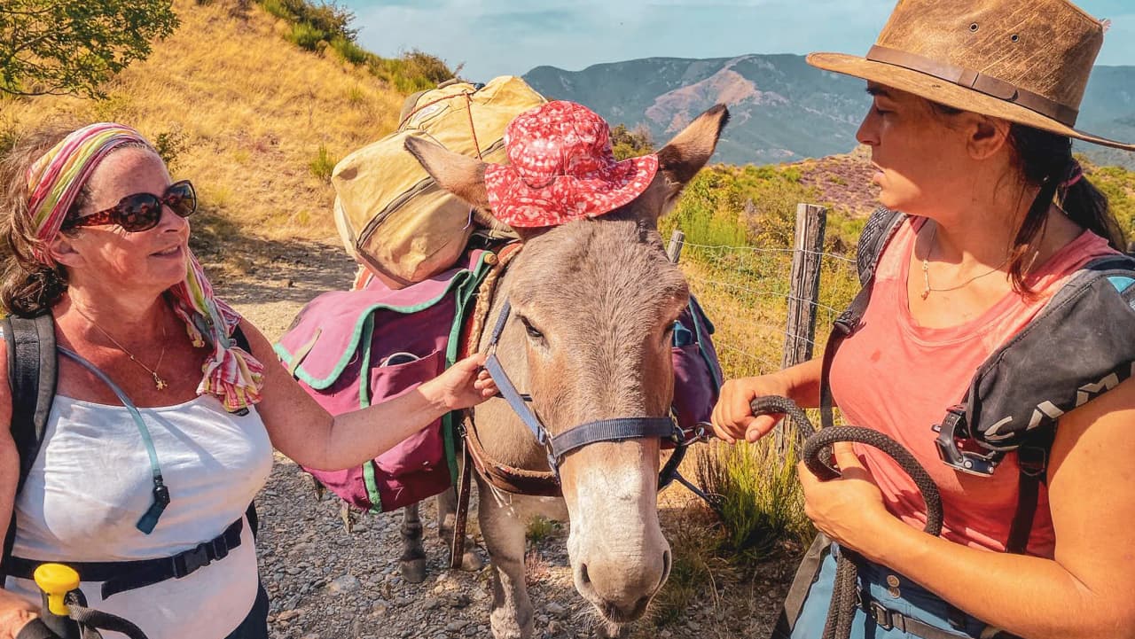 Two smiling hikers, accompanied by a loaded donkey, on a sunny path in the Cévennes.