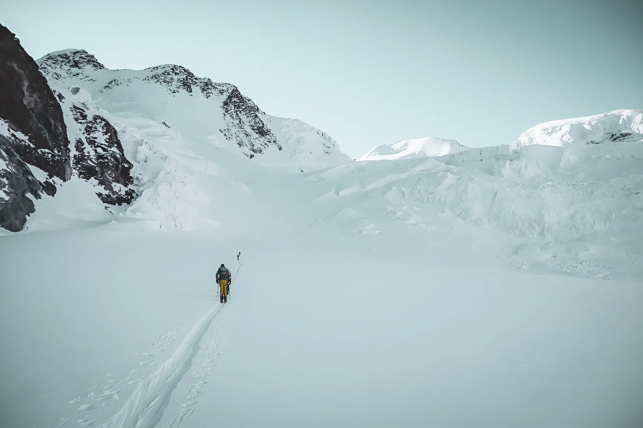Un skieur solitaire traverse un vaste glacier sous un ciel clair, entouré de majestueuses montagnes.