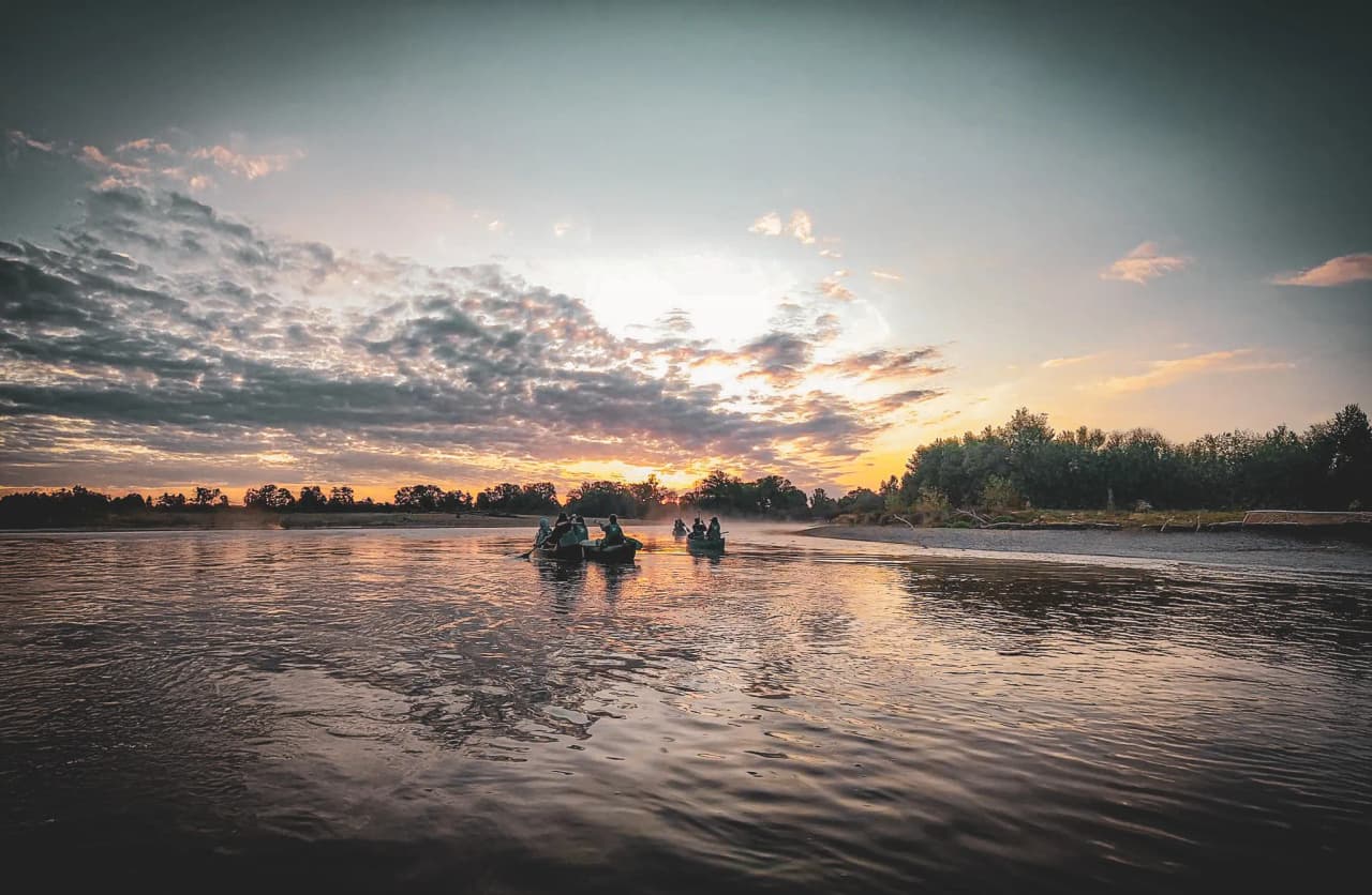 Een vredig landschap langs de rivier, waar verschillende kano's dobberen op het kalme water. De silhouetten van de peddelaars steken af tegen een lucht bezaaid met wolken, getint met oranje en violette tinten.