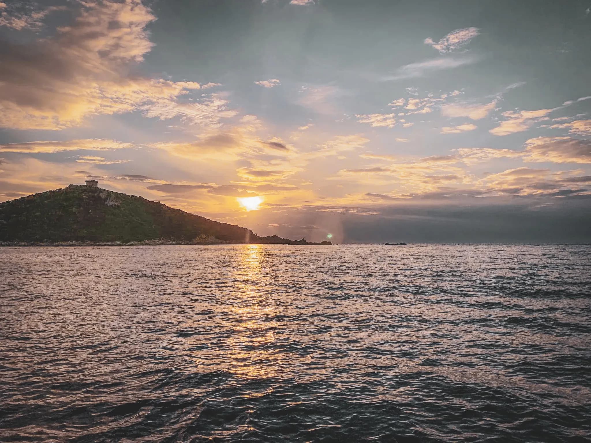 Coucher de soleil doré sur la mer, avec une île verdoyante et une tour génoise en arrière-plan.