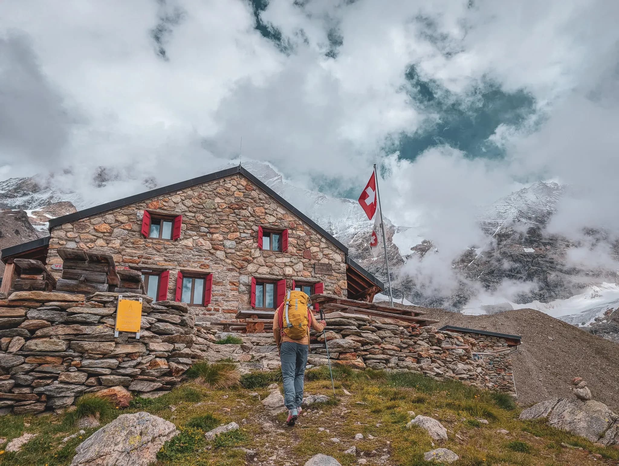 A climber approaches a stone Mountain hut under a cloudy sky, surrounded by majestic mountains.