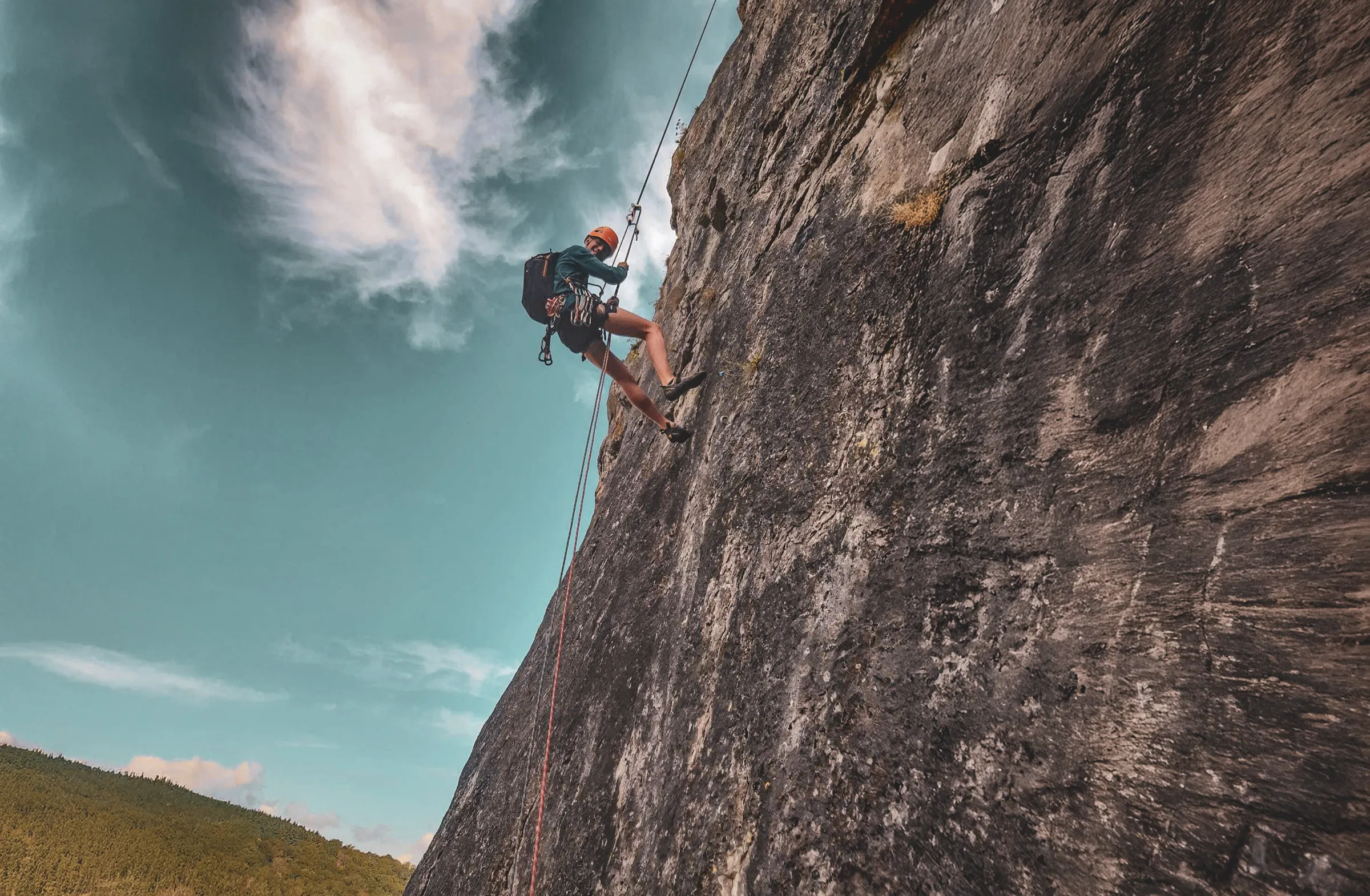 A climber climbing a rock face, using ropes and safety equipment. The sky is a brilliant blue, dotted with bright clouds.