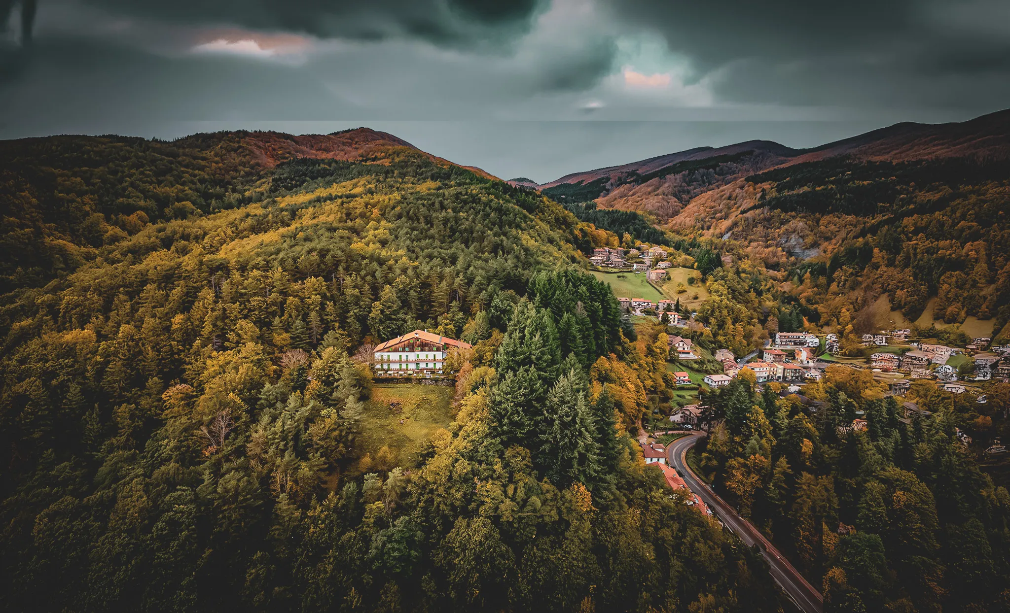 Un paysage montagneux avec une vaste étendue de forêts, principalement de conifères, caractérisé par des couleurs automnales. Au centre, une maison blanche à deux étages se trouve sur une légère pente.