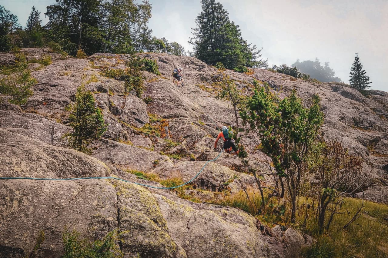 Two mountaineers climb over rocky terrain, surrounded by greenery, facing majestic mountains.
