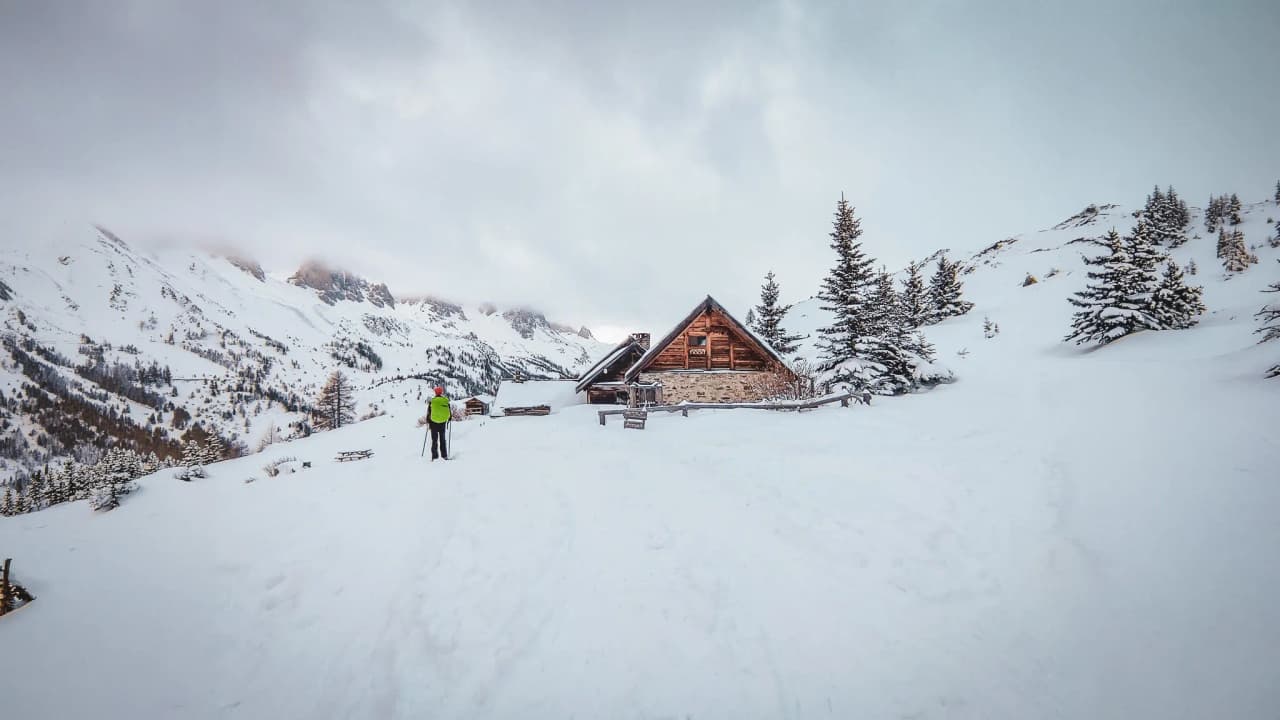 A hiker admires a picturesque mountain hut surrounded by snow and majestic mountains.