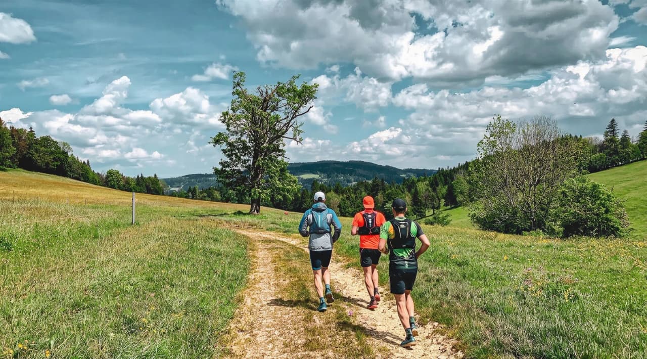 Three runners set off along a green trail in the Jura, under sunny and cloudy skies.