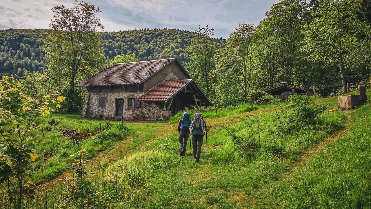 Deux randonneurs cheminent vers une charmante maison en pleine nature, entourée de verdure.