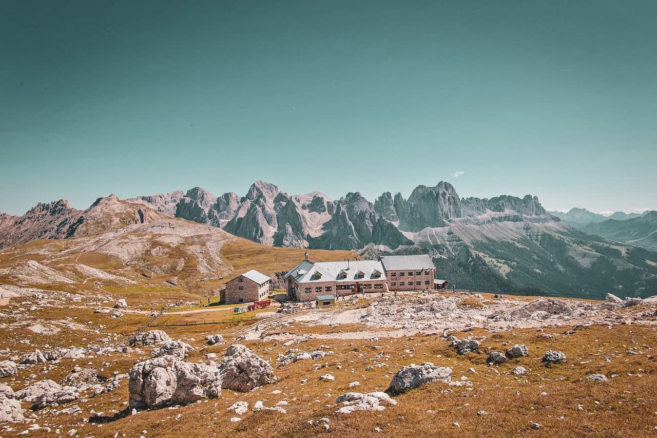 A mountain refuge set in an Alpine landscape, surrounded by vast meadows and rocky outcrops. In the background, a majestic mountain range looms under a clear blue sky. The refuge's stone architecture
