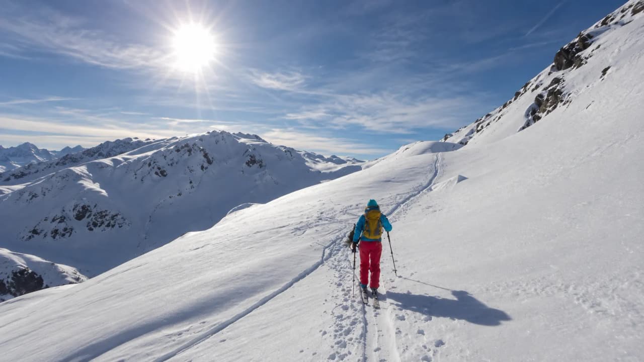 A cross-country skier is making their way down snow-covered slopes under a brilliant blue sky.