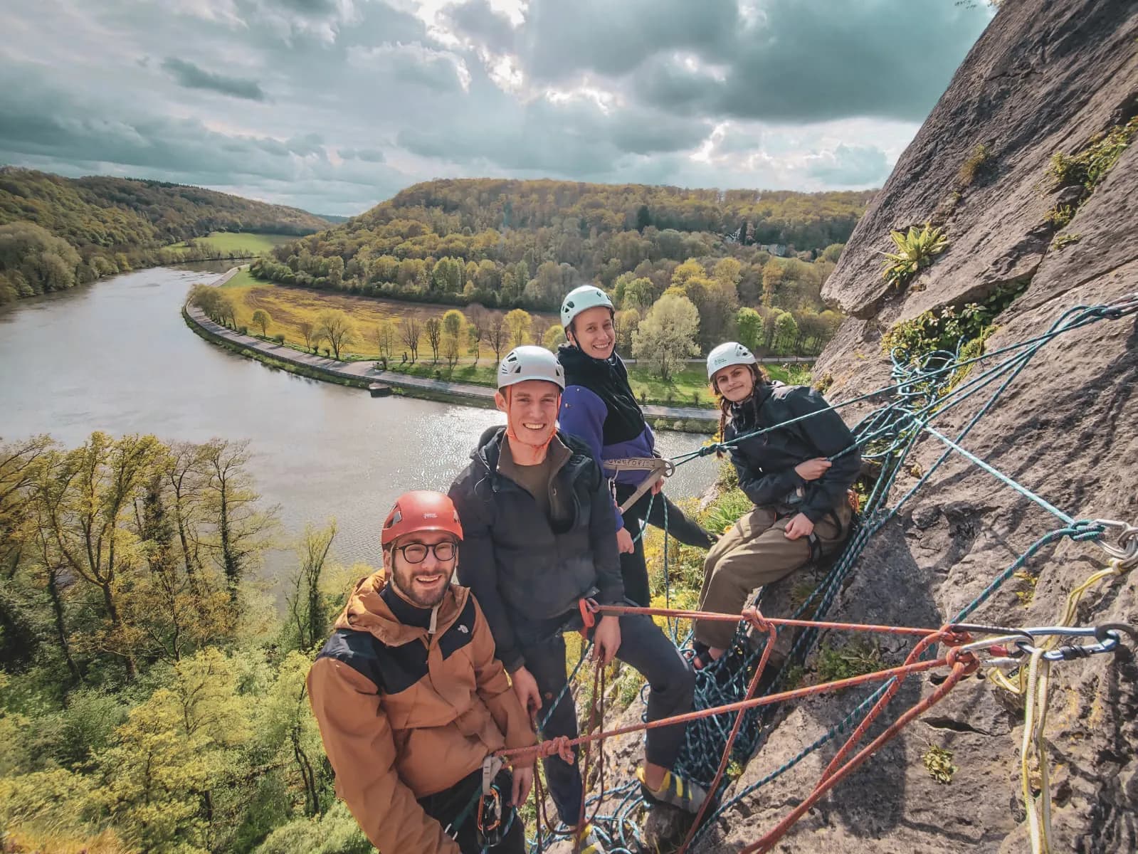 Quatre grimpeurs souriants posent sur une falaise surplombant une belle vallée verdoyante.