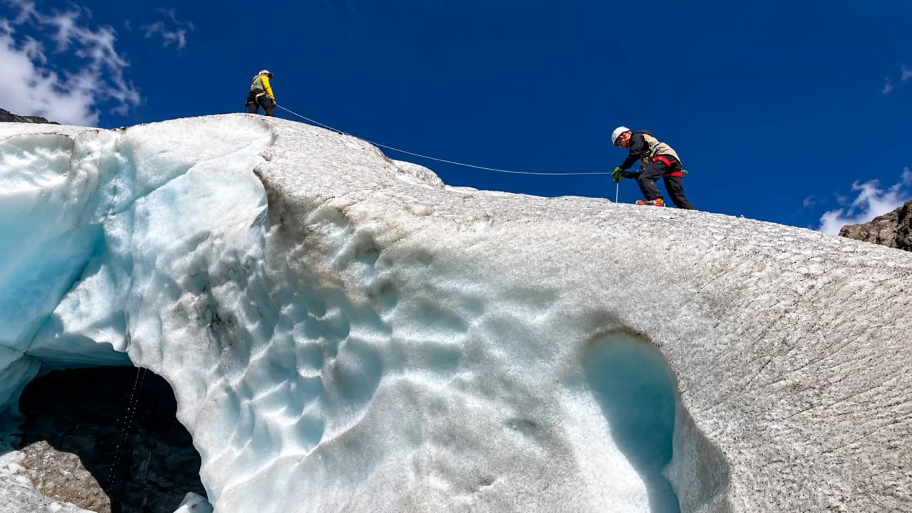 Twee klimmers aan het touw op een ijsbult boven een gletsjergrot, tijdens de gletsjertraining.