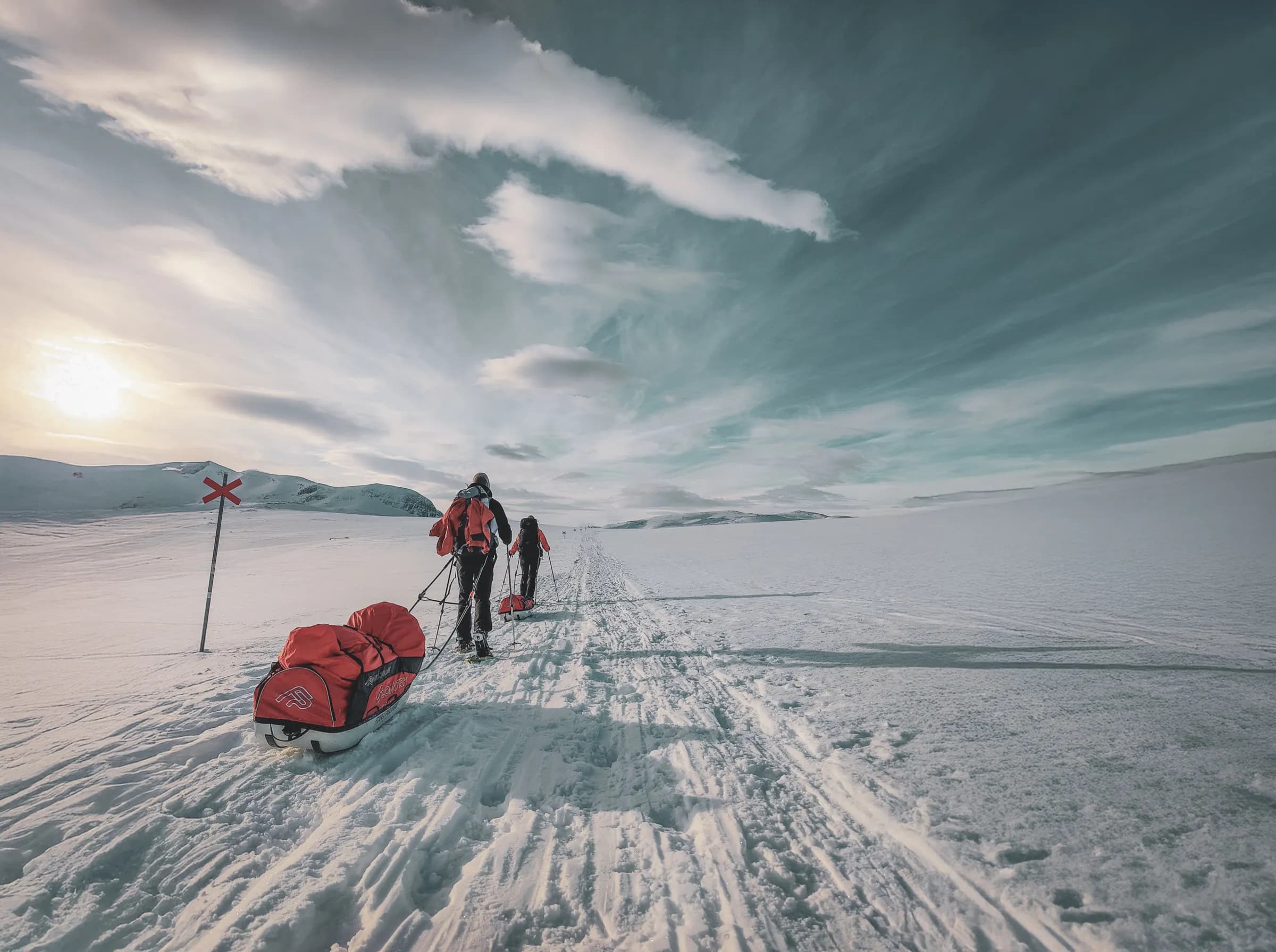 Two hikers on snowshoes pull pulkas across a vast field of snow, under a dramatic sky.