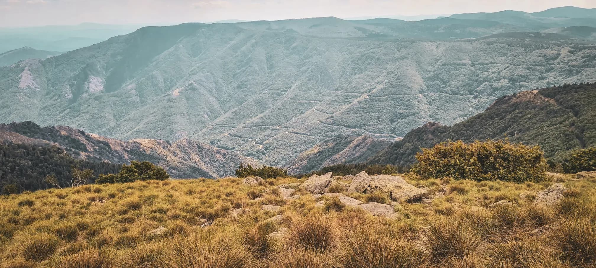 Een uitgestrekt berglandschap van zachte kleuren, bezaaid met rotsen en groen. Een uitnodiging om te ontsnappen.
