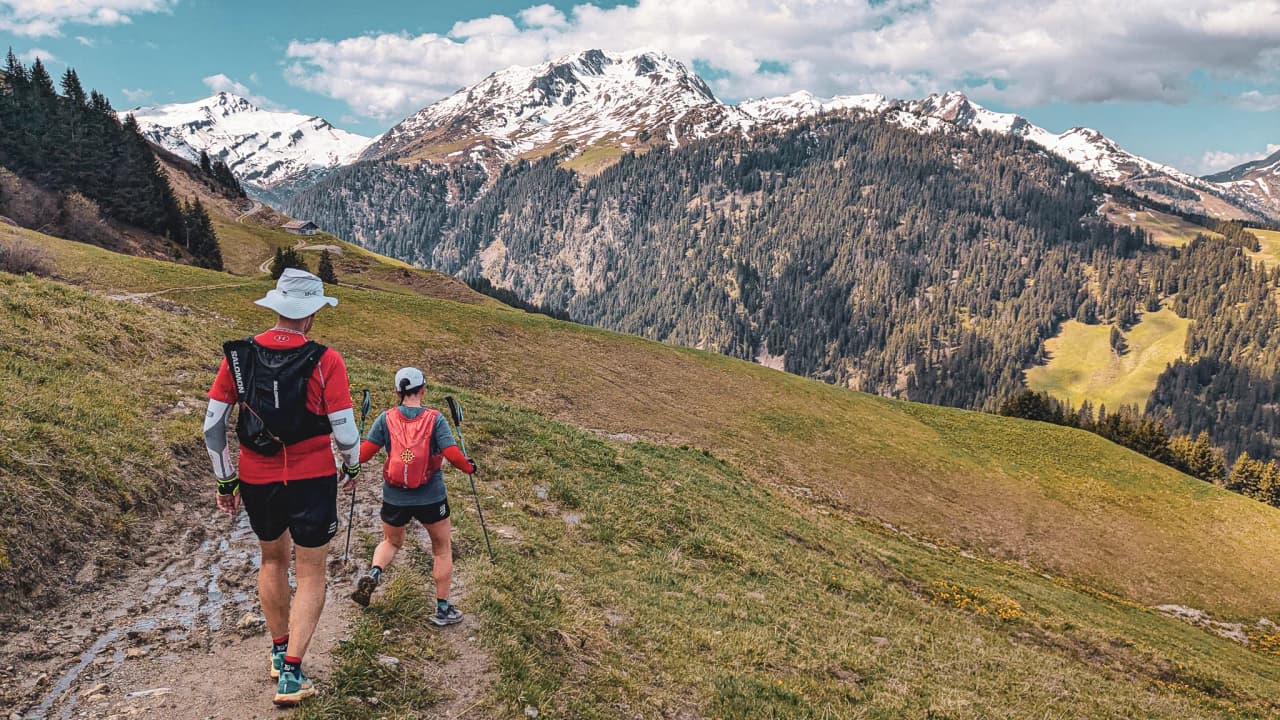 Deux randonneurs sur un sentier verdoyant, avec des montagnes enneigées en arrière-plan.