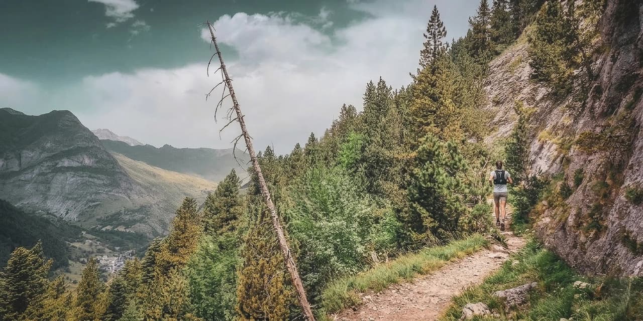 Une coureuse de trail progresse sur un sentier escarpé à flanc de montagne, dominant une vallée boisée et profonde sous un ciel chargé.