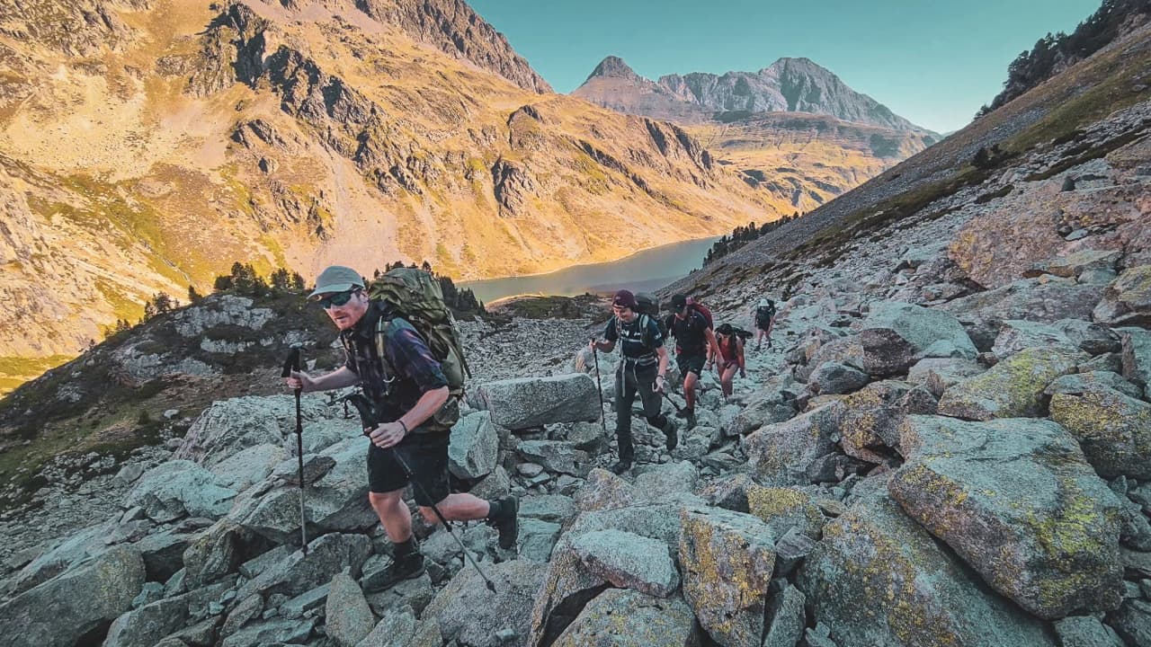 Een groep wandelaars die rotsen beklimt, met berglandschappen en een meer op de achtergrond.