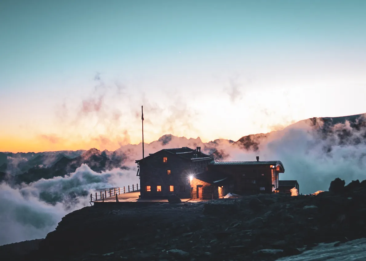 Refuge alpin illuminé au crépuscule, entouré de nuages et montagnes majestueuses, invitation à l'aventure.