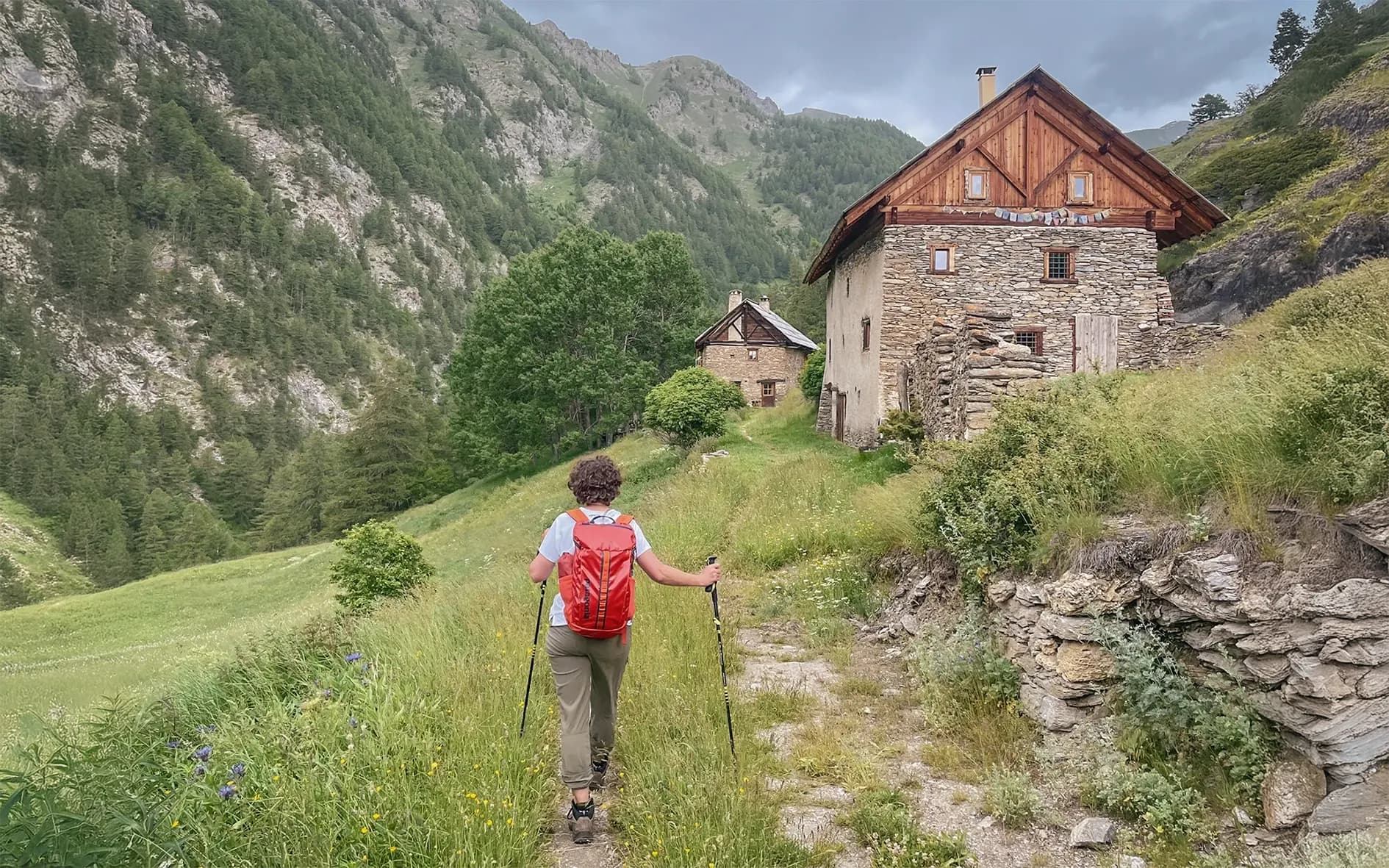A hiker on a green path, surrounded by mountains, near charming old buildings.