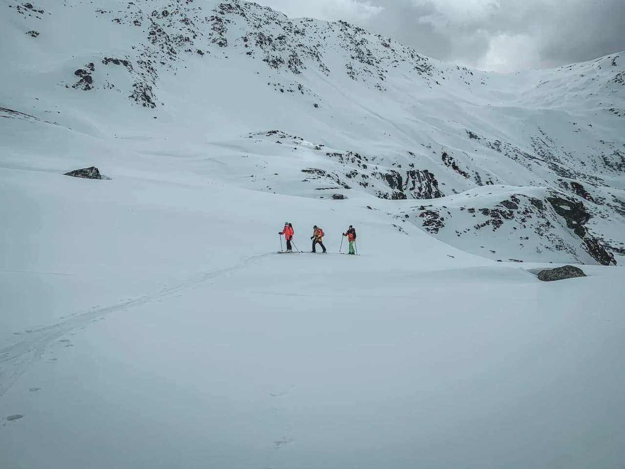 Drie toerskiërs trekken over een uitgestrekt sneeuwplateau onder een bewolkte hemel.
