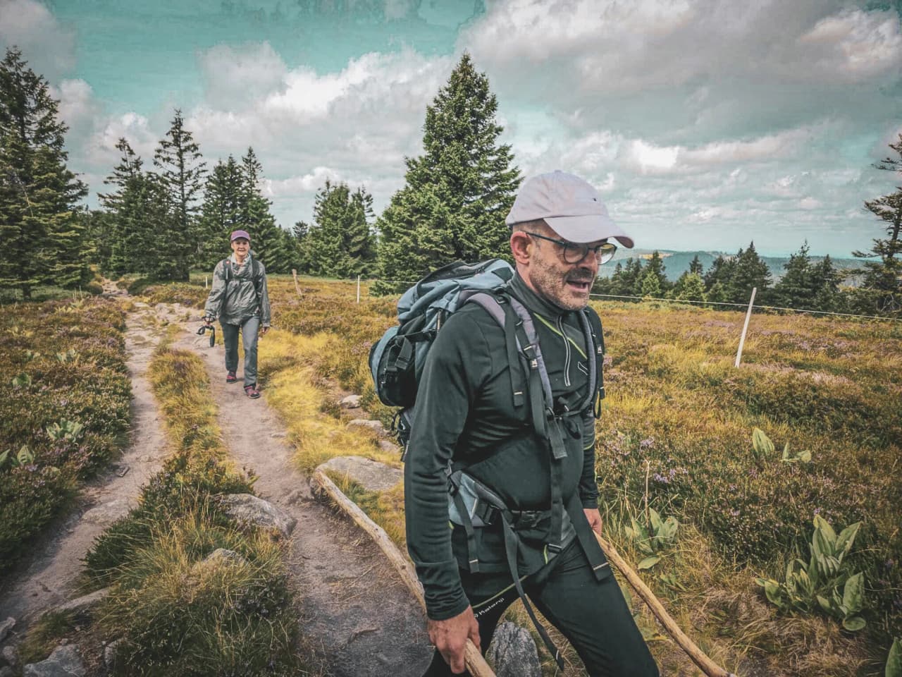 Two hikers make their way along a green path in the Vosges, surrounded by forests and blue skies.