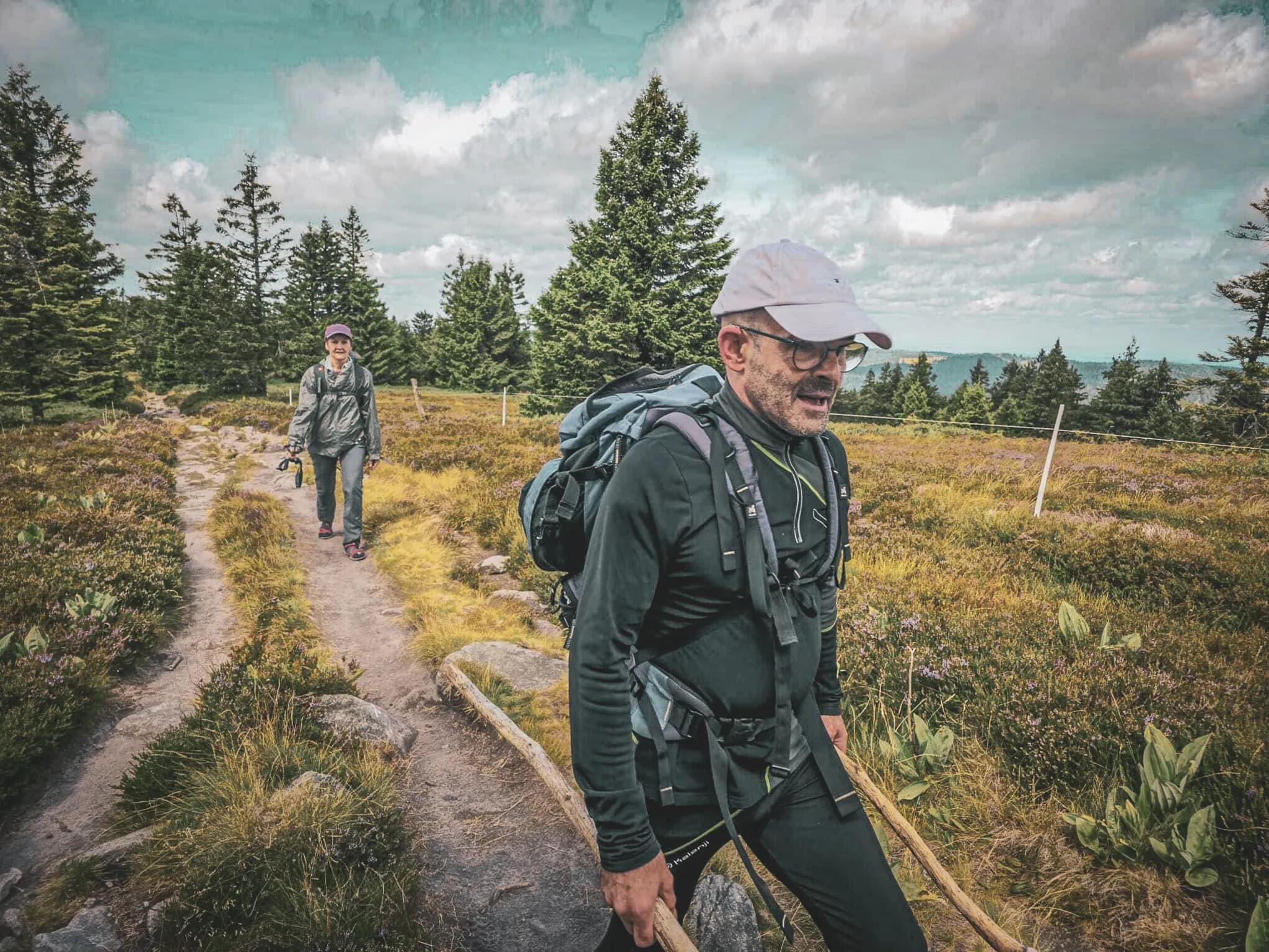 Two hikers make their way along a green path in the Vosges, surrounded by forests and blue skies.