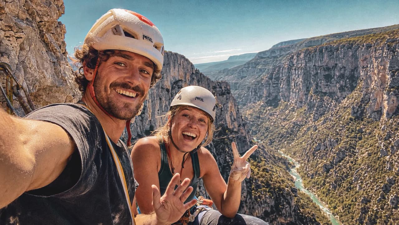Two smiling climbers pose at the top of the Gorges du Verdon, with majestic cliffs in the background.