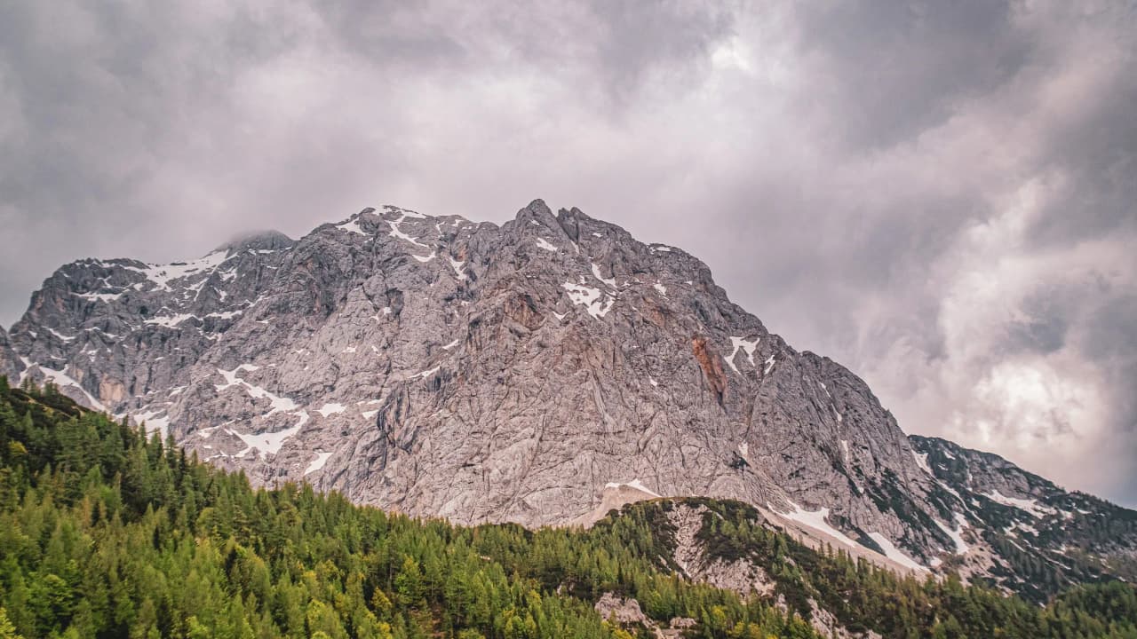 A majestic Alpine peak, adorned with snow, overlooking a verdant forest under a cloudy sky.