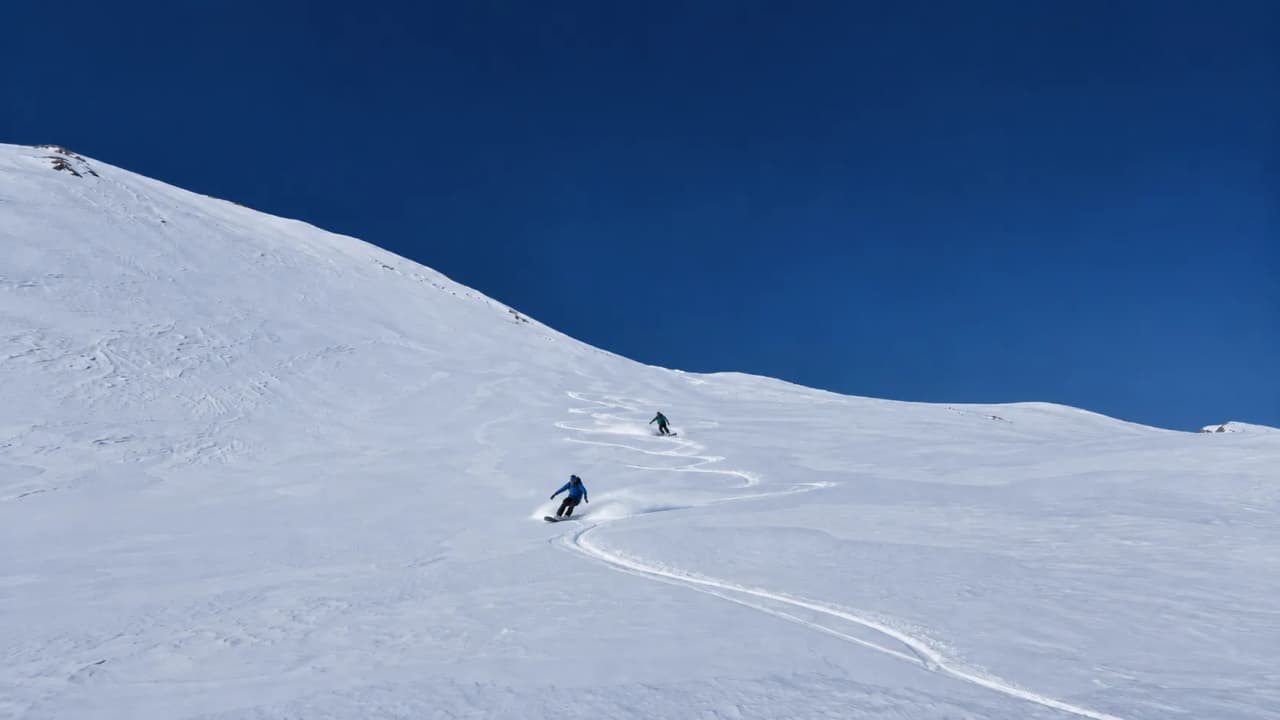 Two snowboarders are riding down a powder-covered slope beneath a clear blue sky in the Clarée Valley.