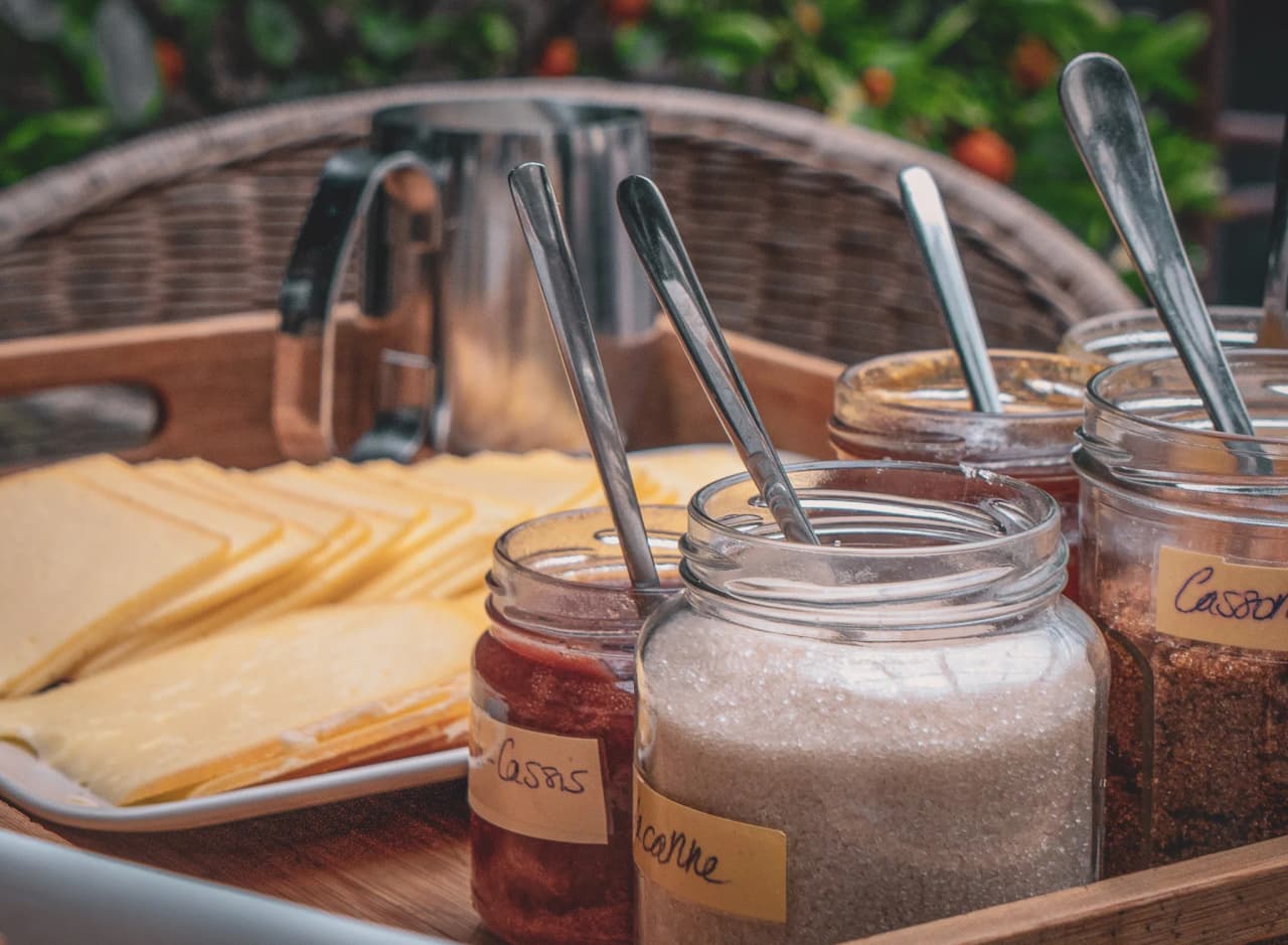Een houten dienblad met plakjes kaas op een wit bord. Verschillende glazen potten zijn gevuld met verschillende substanties: één bevat een mengsel van fruit, een ander suiker, en weer een ander kruiden.