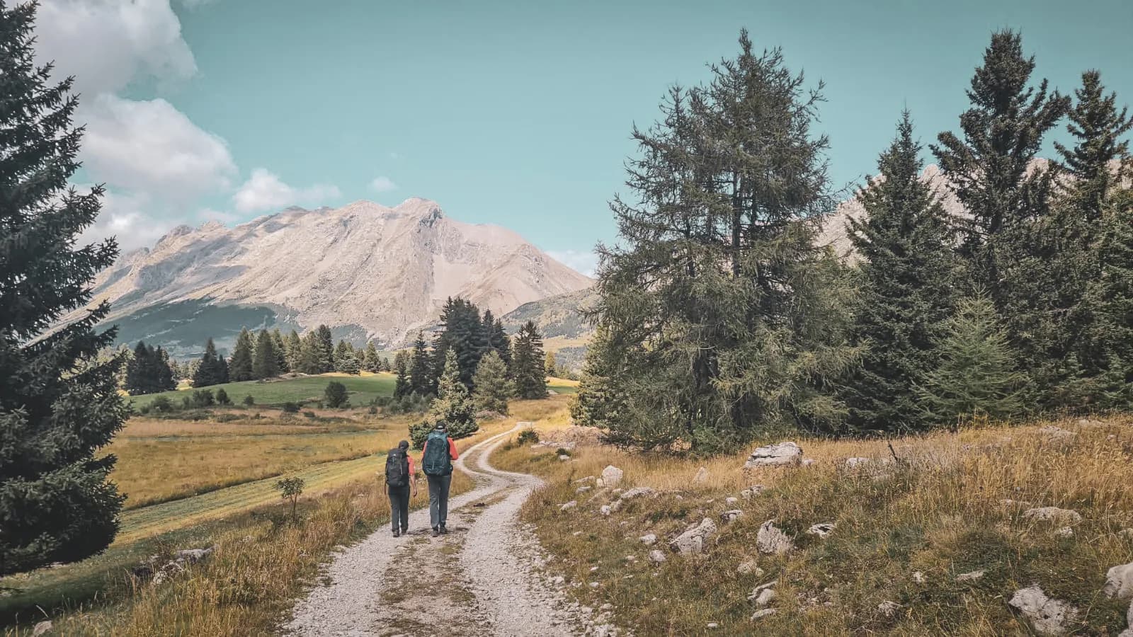 Two hikers walk along a winding path, surrounded by mountains and green forests.