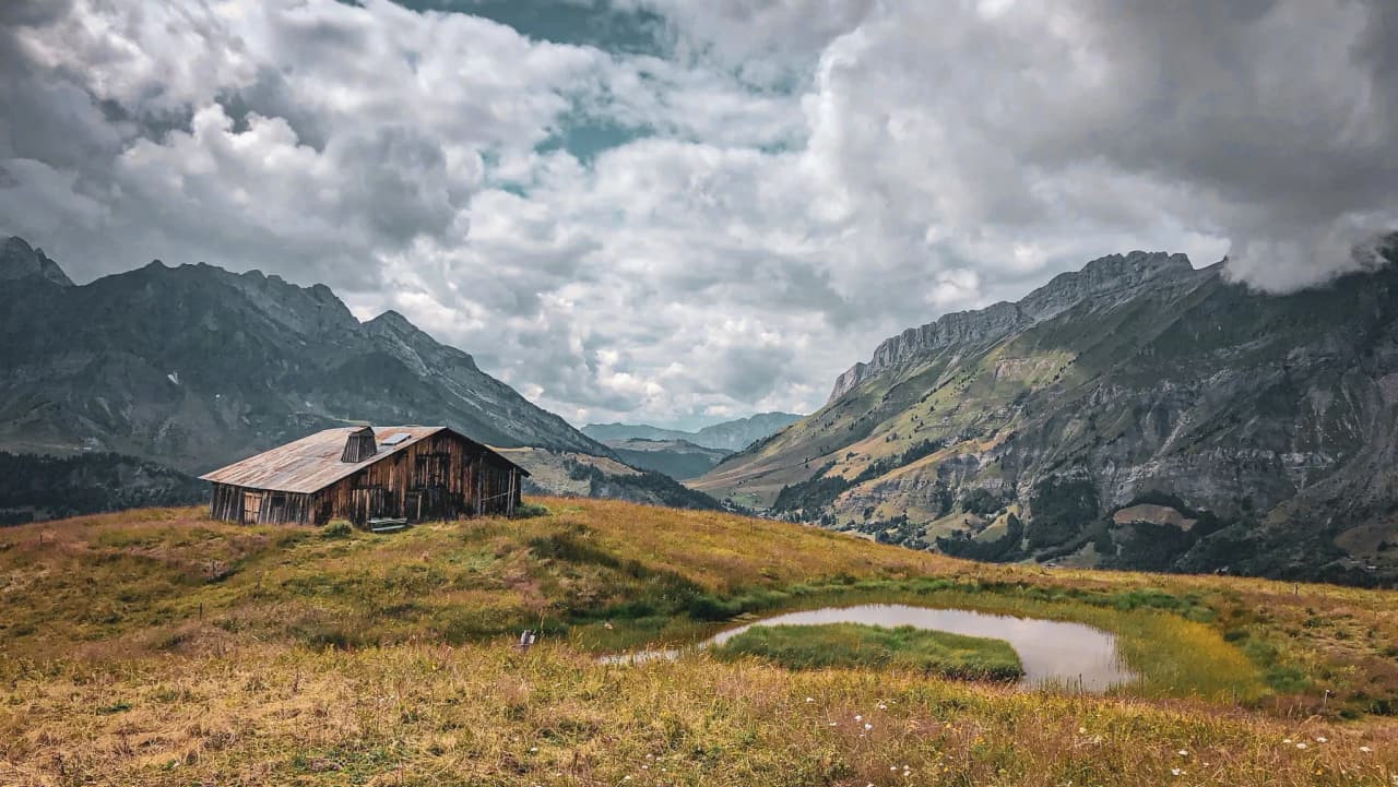 A rustic cabin in the middle of meadows, with majestic mountains in the background.