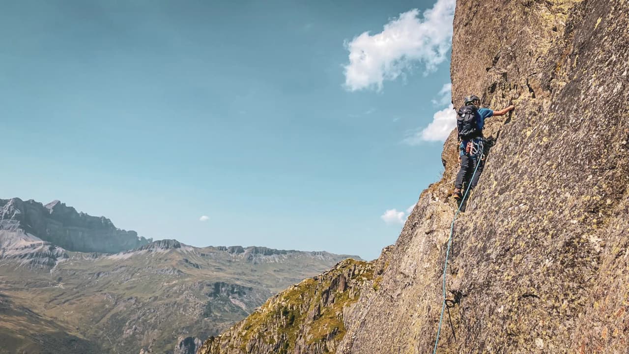 Un alpiniste grimpe une paroi rocheuse sous un ciel bleu, avec des montagnes majestueuses en arrière-plan.