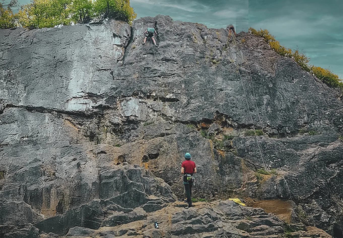A climber is scaling a rocky cliff, while another climber is already at the top, against a backdrop of cloudy skies.