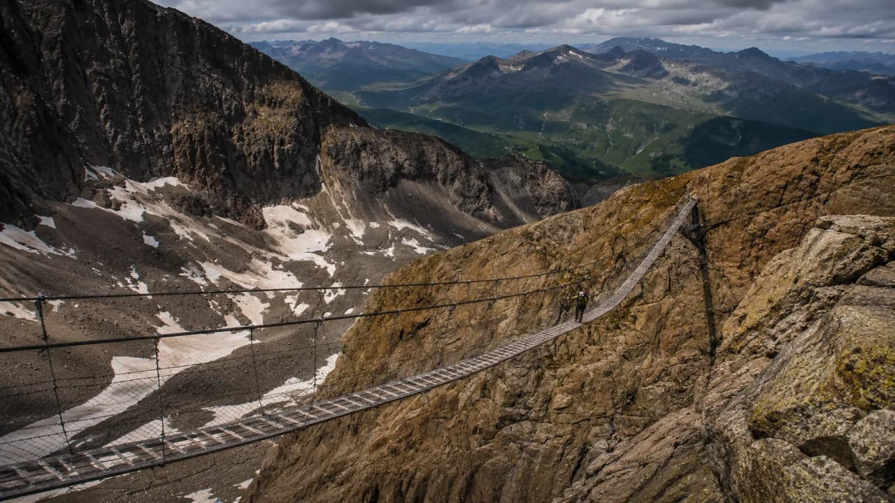 Een wandelaar steekt een hangbrug over majestueuze Alpenlandschappen over.